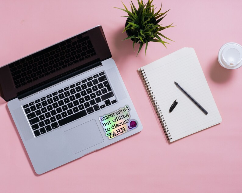 May include: A laptop computer with a sticker that reads "Introverted but willing to discuss YARN."  A notebook with a pen and a cup of coffee are on a pink surface.