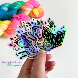 May include: A set of iridescent holographic stickers featuring a black and white illustration of a book, yarn, and knitting needles. The stickers are arranged in a fan shape and have a rainbow iridescent finish. The text "Green Fox Farms Designs" is visible in the bottom left corner of the image.