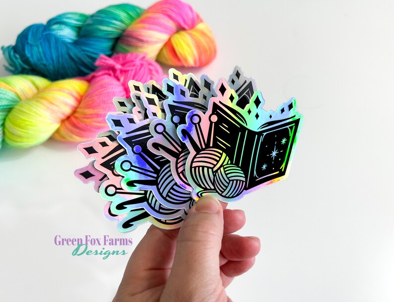 May include: A set of iridescent holographic stickers featuring a black and white illustration of a book, yarn, and knitting needles. The stickers are arranged in a fan shape and have a rainbow iridescent finish. The text "Green Fox Farms Designs" is visible in the bottom left corner of the image.