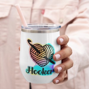 May include: White stainless steel wine tumbler with a colourful yarn ball and crochet hook design and the text "Hooker" in a rainbow font.
