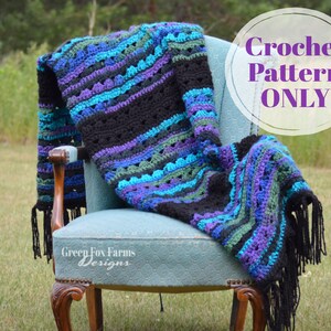 Op de afbeelding: Een handgemaakte gehaakte deken in tinten zwart, teal, paars en groen, gedrapeerd over een vintage blauwe stoel. De deken heeft een gestreept patroon en franjes. De tekst "Crochet Pattern ONLY" staat in een witte cirkel.