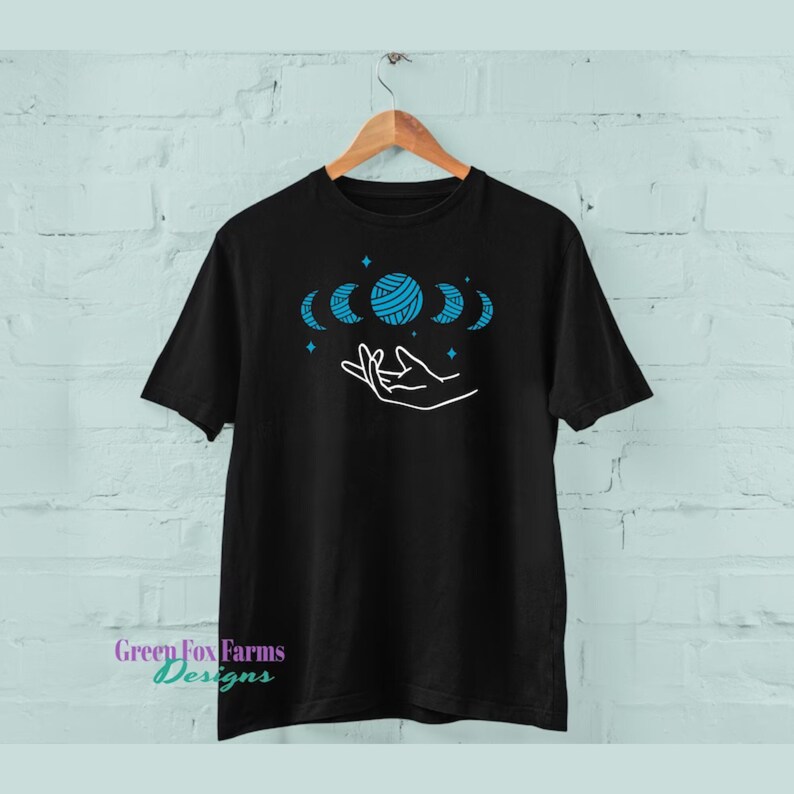 May include: Black t-shirt with a white outline of a hand holding a blue yarn ball with crescent moons around it. The yarn ball is made of a textured pattern. There are small white stars scattered around the design.