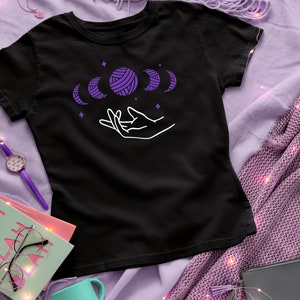 May include: Black t-shirt with a white outline of a hand holding a purple yarn ball with a crescent moon design. The design is surrounded by stars.