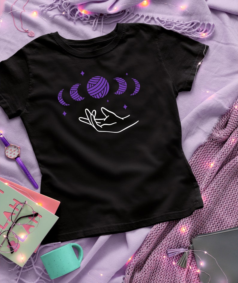 May include: Black t-shirt with a white outline of a hand holding a purple yarn ball with a crescent moon design. The design is surrounded by stars.