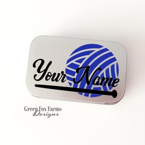 May include: A silver metal tin with a blue and white yarn ball and a black knitting needle graphic. The text "Your Name" is printed in black cursive font. The text "Green Fox Farms Designs" is printed in gray below the graphic.