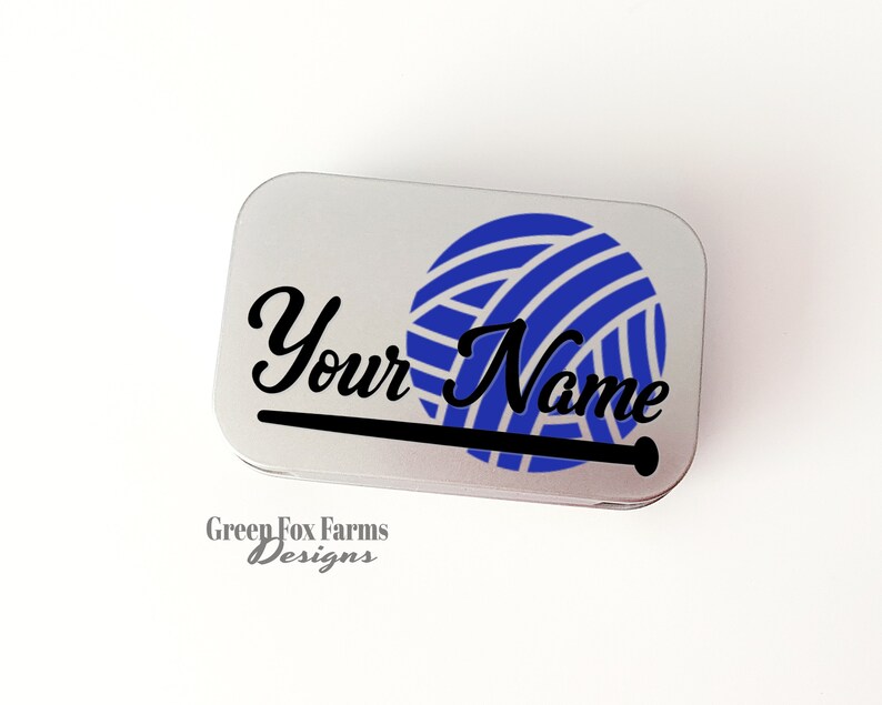 May include: A silver metal tin with a blue and white yarn ball and a black knitting needle graphic. The text "Your Name" is printed in black cursive font. The text "Green Fox Farms Designs" is printed in gray below the graphic.