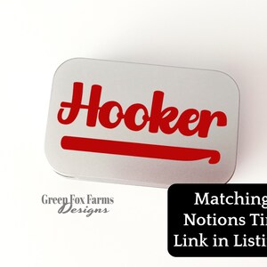 May include: A silver metal tin with the word "Hooker" in red lettering and a red crochet hook graphic. The tin is labelled "Green Fox Farms Designs".