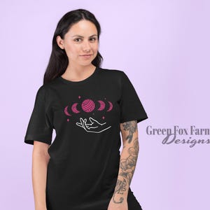 May include: A black t-shirt with a pink yarn ball and crescent moon design. The design features a hand holding the yarn ball and moons. The text "Green Fox Farms Designs" is visible in the bottom right corner.