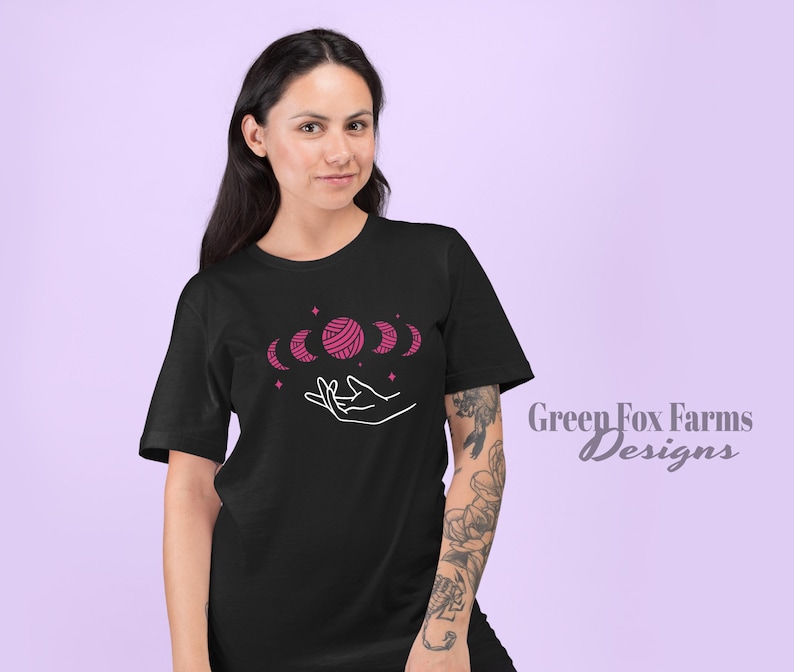 May include: A black t-shirt with a pink yarn ball and crescent moon design. The design features a hand holding the yarn ball and moons. The text "Green Fox Farms Designs" is visible in the bottom right corner.