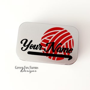 May include: A silver metal tin with a red and white yarn ball and a black crochet hook. The text "Your Name" is written in black cursive lettering on the tin.