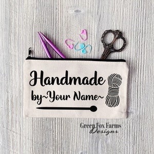May include: A white canvas zippered pouch with black text that reads "Handmade by~Your Name~" and a black and white illustration of a ball of yarn. The pouch has a black zipper and a black line across the bottom. The text "Green Fox Farms Designs" is printed in black at the bottom of the pouch.