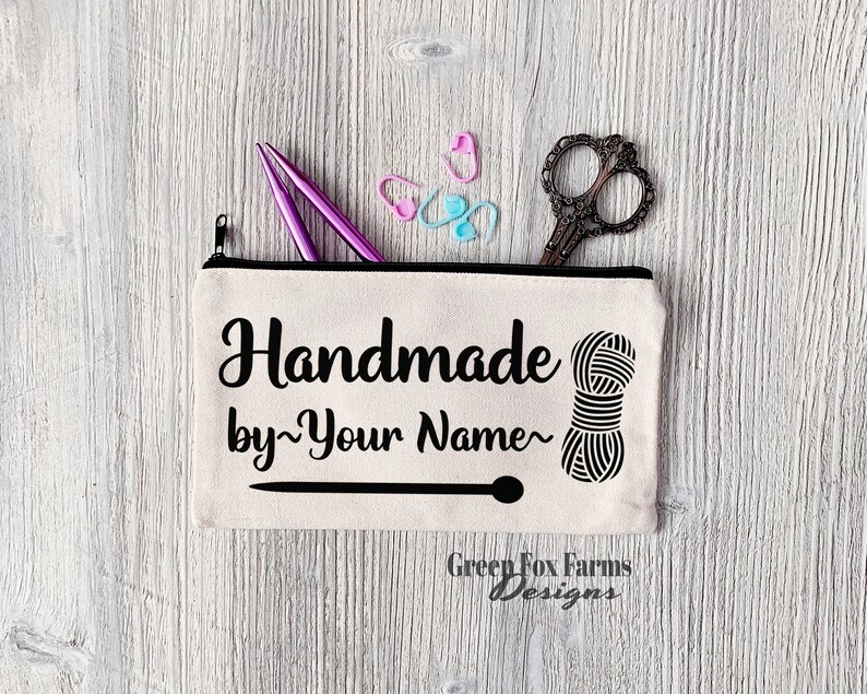 May include: A white canvas zippered pouch with black text that reads "Handmade by~Your Name~" and a black and white illustration of a ball of yarn. The pouch has a black zipper and a black line across the bottom. The text "Green Fox Farms Designs" is printed in black at the bottom of the pouch.