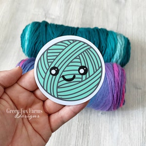 May include: A teal blue ball of yarn with a cute cartoon face, smiling with big eyes. The yarn ball is on a white background.