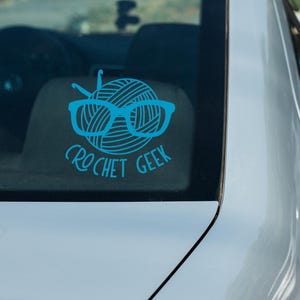 May include: A blue car window sticker with a ball of yarn wearing glasses and the text "crochet geek".