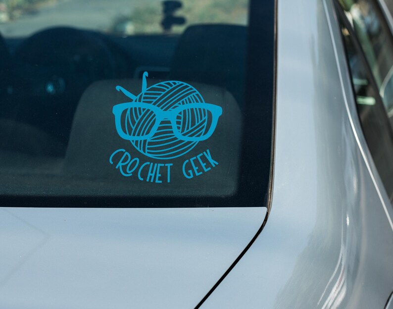 May include: A blue car window sticker with a ball of yarn wearing glasses and the text "crochet geek".