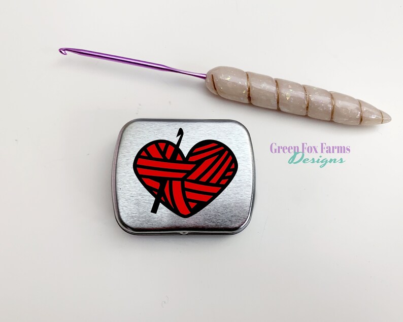 Puede incluir: Una lata de metal plateada con un dise&ntilde;o de coraz&oacute;n rojo que presenta un ovillo de lana y un gancho de ganchillo. El texto "Green Fox Farms Designs" est&aacute; impreso en la lata.