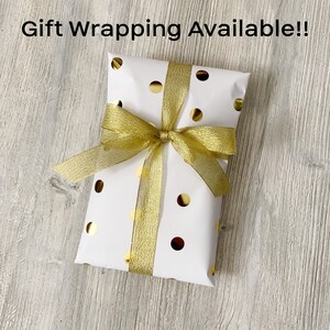 May include: A white gift box with gold polka dots and a gold ribbon tied in a bow. The box is sitting on a wooden surface. The text "Gift Wrapping Available!" is written above the box.
