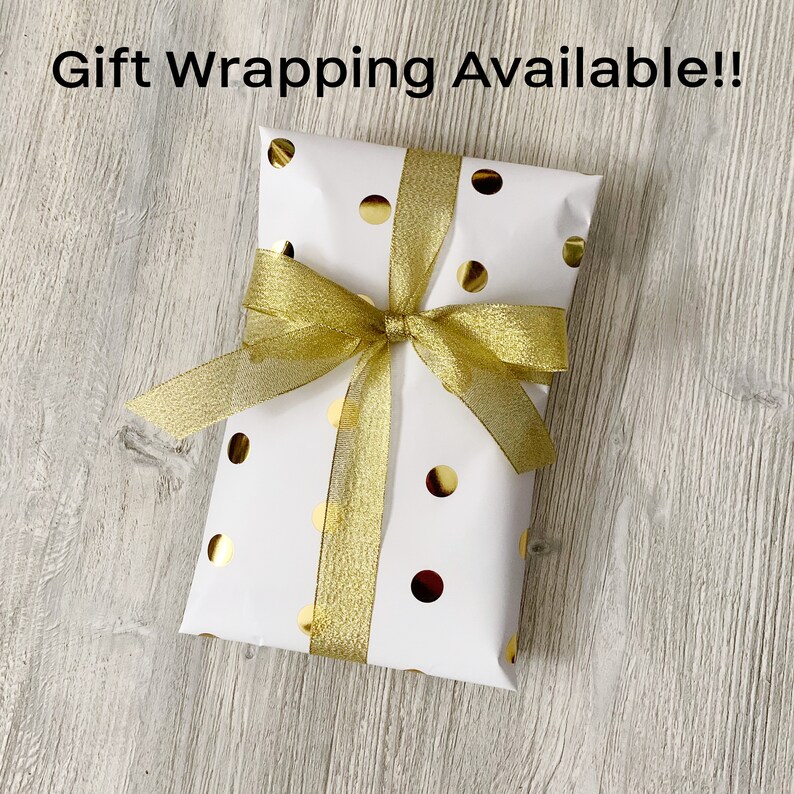 May include: A white gift box with gold polka dots and a gold ribbon tied in a bow. The box is sitting on a wooden surface. The text "Gift Wrapping Available!" is written above the box.
