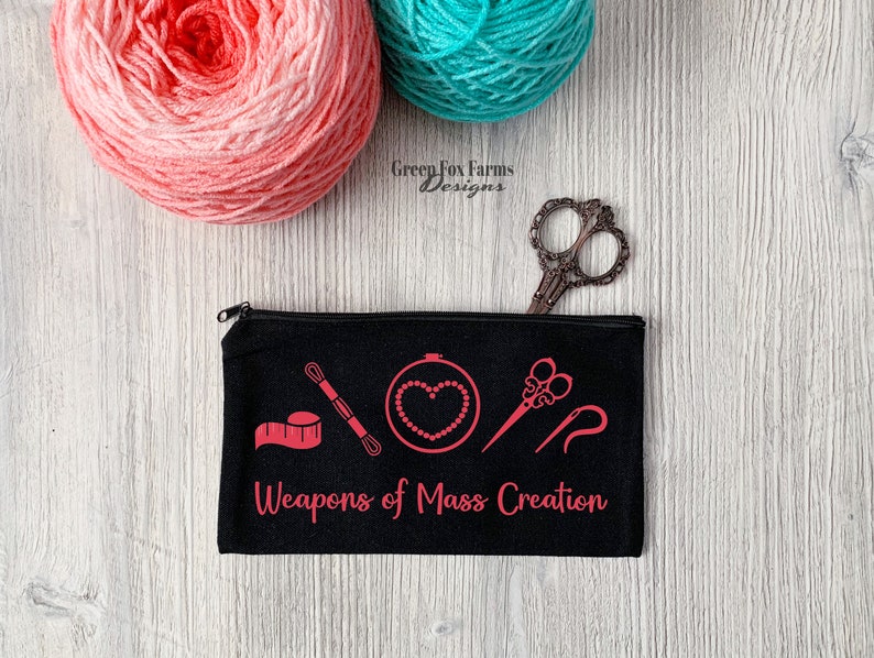 May include: Black zippered pouch with the text "Weapons of Mass Creation" in pink. The pouch has a pink design of sewing tools, including a needle, thread, scissors, and a tape measure.