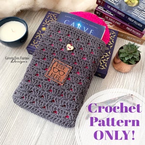 May include: A gray crochet book cover with a pink border and a brown leather patch that says "Book Nerd" with a pair of glasses on it. The cover is sitting on a wooden surface with a stack of books and a small potted plant.