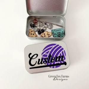 May include: A silver metal tin with a purple yarn ball and the word "Custom" in black lettering. The tin has the words "Green Fox Farms Designs" printed on the bottom.