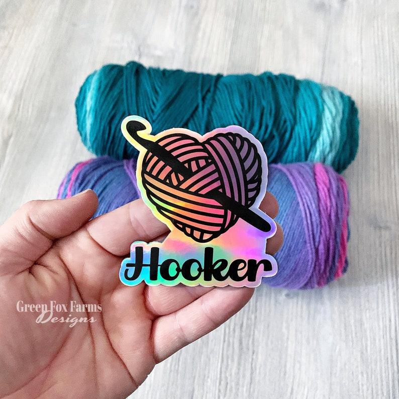 May include: A holographic sticker featuring a heart-shaped ball of yarn and a crochet hook, with the word "Hooker" written beneath. The sticker is held in a hand, against a backdrop of teal and purple yarn.