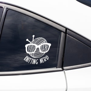 May include: White vinyl sticker on a car window with a ball of yarn wearing glasses and the text "Knitting Nerd".