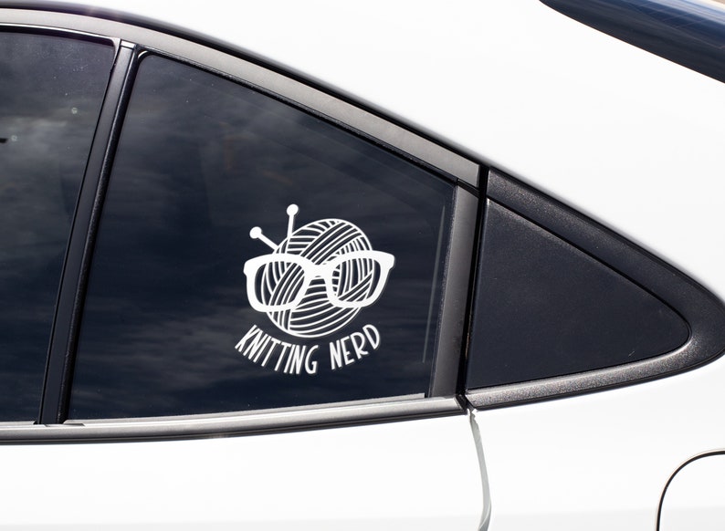 May include: White vinyl sticker on a car window with a ball of yarn wearing glasses and the text "Knitting Nerd".