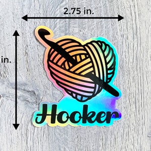 May include: A holographic sticker featuring a rainbow yarn ball and crochet hook, with the word "Hooker" in black text.