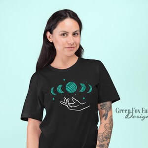 May include: Black t-shirt with a design of a hand holding five moons, one of which is a ball of yarn. The moons are teal blue and the design is outlined in white. The text "Green Fox Farms Designs" is in the bottom right corner.