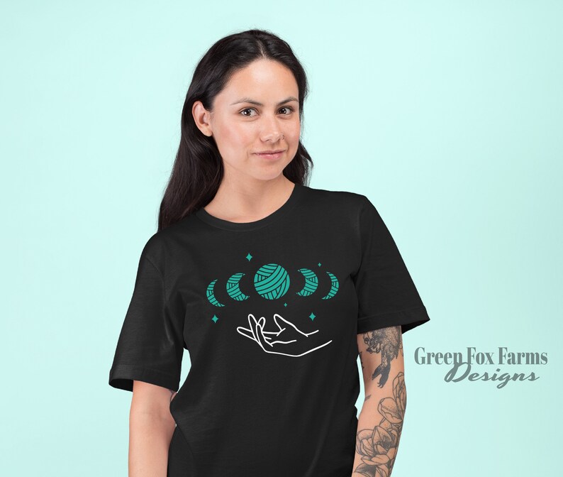 May include: Black t-shirt with a design of a hand holding five moons, one of which is a ball of yarn. The moons are teal blue and the design is outlined in white. The text "Green Fox Farms Designs" is in the bottom right corner.