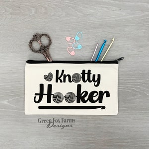 May include: A white canvas zippered pouch with the text "Knotty Hooker" in black lettering. The pouch has a black zipper and a black hook design. The text "Green Fox Farms Designs" is printed in black at the bottom of the pouch.