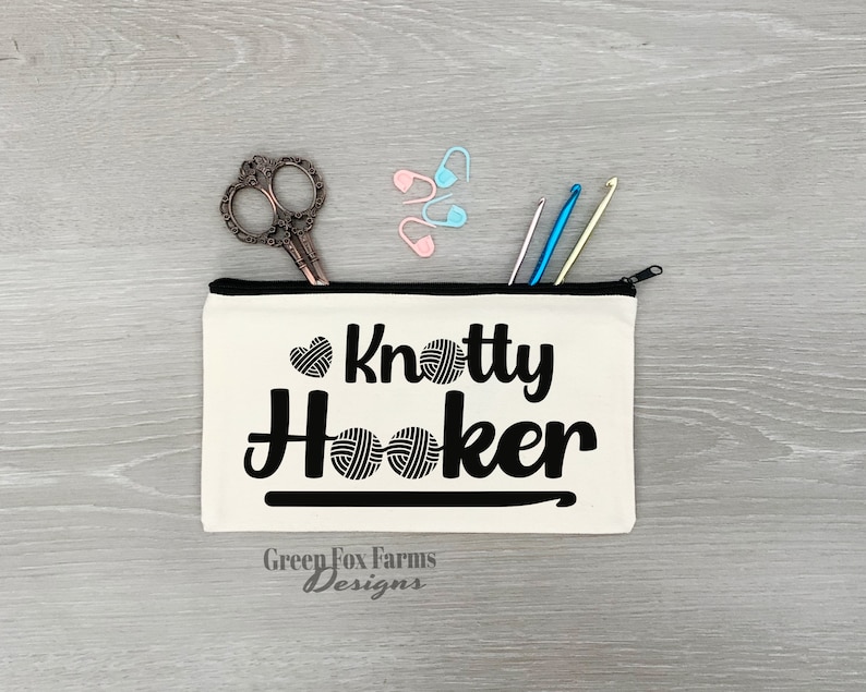May include: A white canvas zippered pouch with the text "Knotty Hooker" in black lettering. The pouch has a black zipper and a black hook design. The text "Green Fox Farms Designs" is printed in black at the bottom of the pouch.