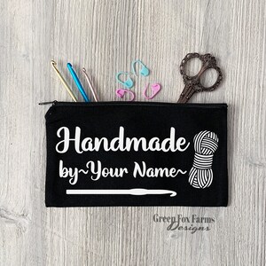 May include: Black canvas zippered pouch with white text that reads "Handmade by~Your Name~" and a white yarn ball and crochet hook graphic. The pouch is on a wood background with crochet hooks, stitch markers, and scissors.