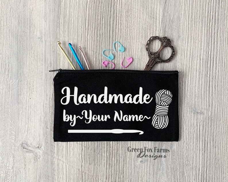 May include: Black canvas zippered pouch with white text that reads "Handmade by~Your Name~" and a white yarn ball and crochet hook graphic. The pouch is on a wood background with crochet hooks, stitch markers, and scissors.