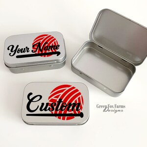 May include: Two silver metal tins with a red and white yarn ball and knitting needle design. One tin says "Your Name" and the other says "Custom". The bottom tin has the text "Green Fox Farms Designs".