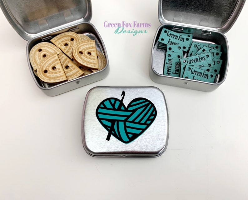 Puede incluir: Dos latas de metal plateadas con tapas. La lata de la izquierda contiene botones de madera en forma de coraz&oacute;n. La lata de la derecha contiene peque&ntilde;as etiquetas azules con el texto "Green Fox Farms". La lata del centro tiene un gr&aacute;fico de una madeja de hilo turquesa con agujas de tejer.