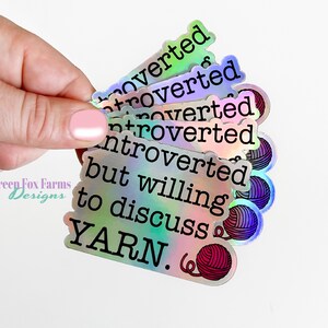 May include: A stack of iridescent holographic stickers with the text "Introverted but willing to discuss YARN."  The stickers feature a small yarn ball graphic.
