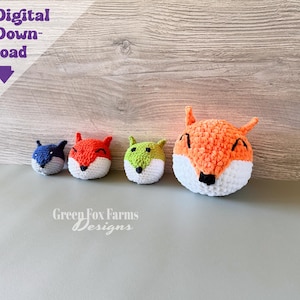 May include: Four crocheted animal heads in varying sizes and colors, including a small blue, a small orange, a small green, and a large orange fox head. Each features embroidered eyes and a nose. The text Digital Download is visible in the corner.