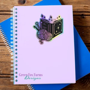 May include: A holographic sticker featuring a black and white illustration of a book with a star and yarn balls. The sticker is on a purple notebook with the text "Green Fox Farms Designs".