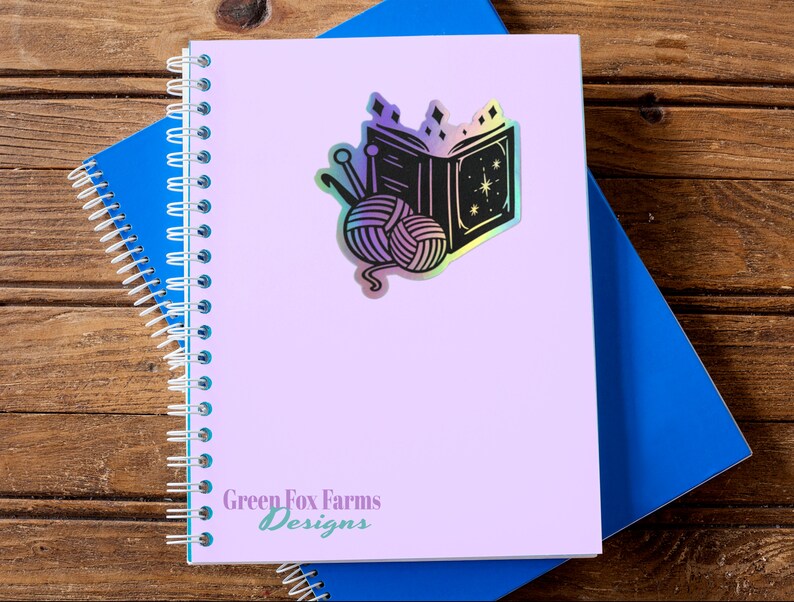 May include: A holographic sticker featuring a black and white illustration of a book with a star and yarn balls. The sticker is on a purple notebook with the text "Green Fox Farms Designs".