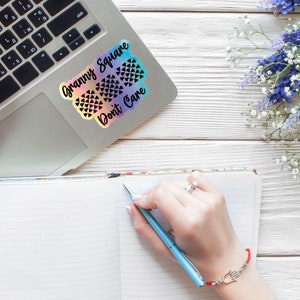 May include: A laptop computer with a sticker that says "Granny Square Don't Care" on the lid. A notebook with lined paper is open on a white wooden surface. A person is writing in the notebook with a blue pen. A bouquet of purple and white flowers is in the upper right corner of the image.