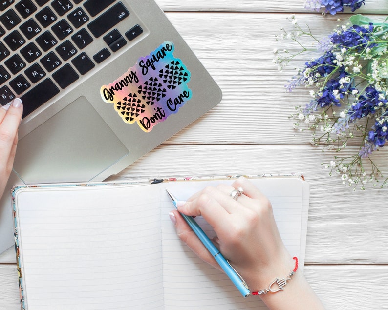 May include: A laptop computer with a sticker that says "Granny Square Don't Care" on the lid. A notebook with lined paper is open on a white wooden surface. A person is writing in the notebook with a blue pen. A bouquet of purple and white flowers is in the upper right corner of the image.