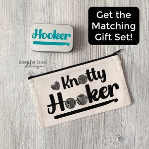 May include: A white canvas zippered pouch with the text "Knotty Hooker" in black lettering. The pouch has a black hook and yarn ball graphic. A silver tin with the word "Hooker" in teal lettering is also shown.