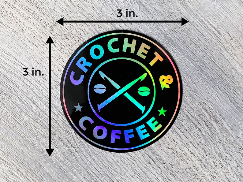 May include: A 3-inch round sticker with a black background and a holographic finish. The sticker features a white design of crossed crochet hooks with coffee beans, surrounded by the text "CROCHET & COFFEE".