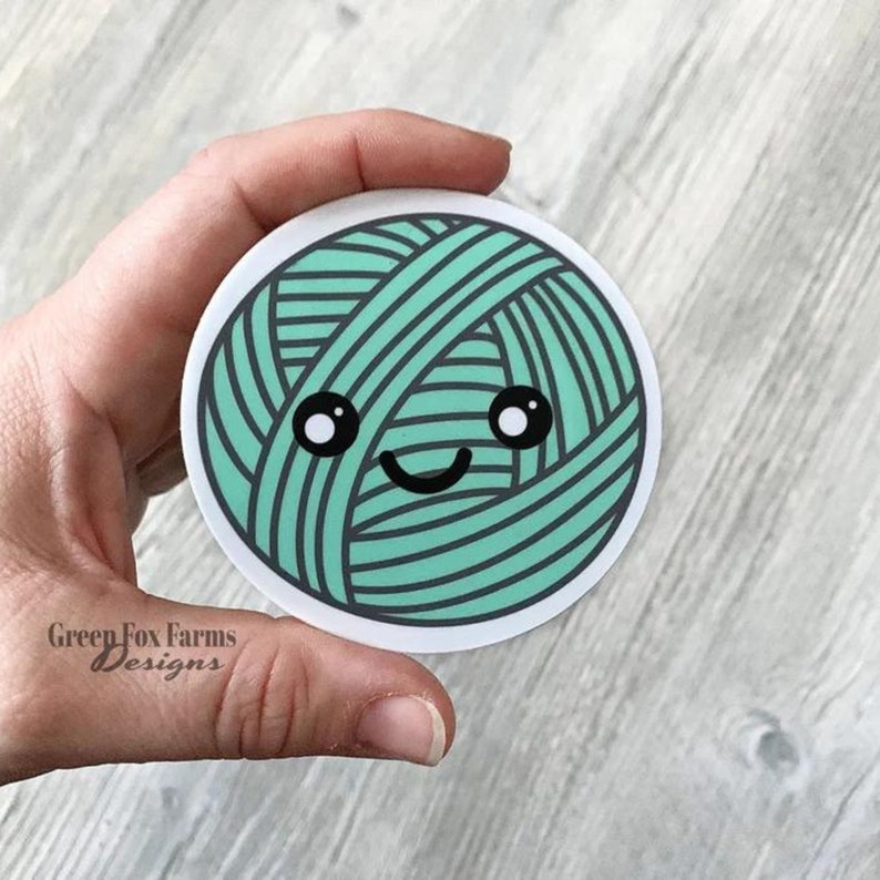 May include: A round sticker featuring a green ball of yarn with a smiling face. The yarn ball has a white background and black eyes.
