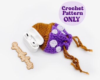 May include: A crochet mushroom-shaped AirPod case in brown and purple with white accents. The case has a drawstring closure and is next to a pair of white AirPods and a wooden sign that says "greenfoxfarms". The text "Crochet Pattern ONLY" is in a purple circle.