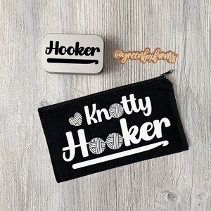 May include: A black canvas zippered pouch with the text "Knotty Hooker" in white lettering. The pouch has a white yarn ball graphic and a white crochet hook graphic. The pouch is on a wood surface.