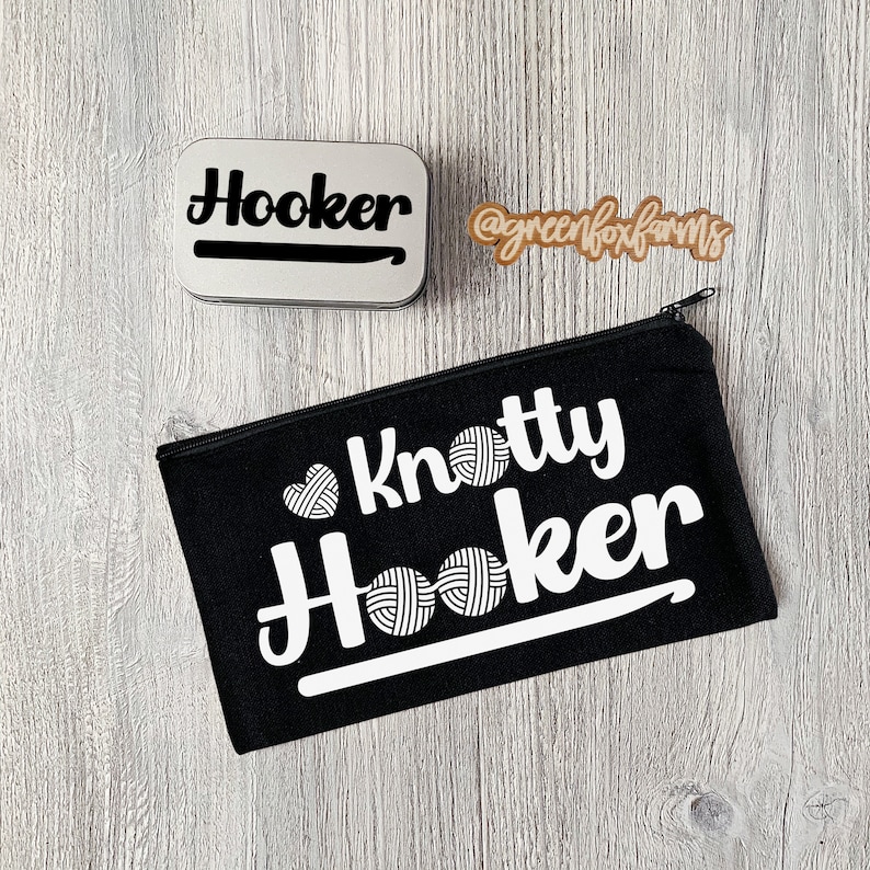 May include: A black canvas zippered pouch with the text "Knotty Hooker" in white lettering. The pouch has a white yarn ball graphic and a white crochet hook graphic. The pouch is on a wood surface.