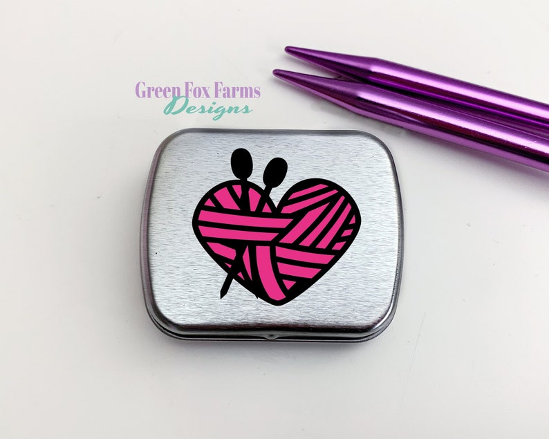Puede incluir: Una lata de metal plateada con un dise&ntilde;o de coraz&oacute;n rosa y negro que presenta un ovillo de lana y agujas de tejer. El texto "Green Fox Farms Designs" est&aacute; impreso en la lata.